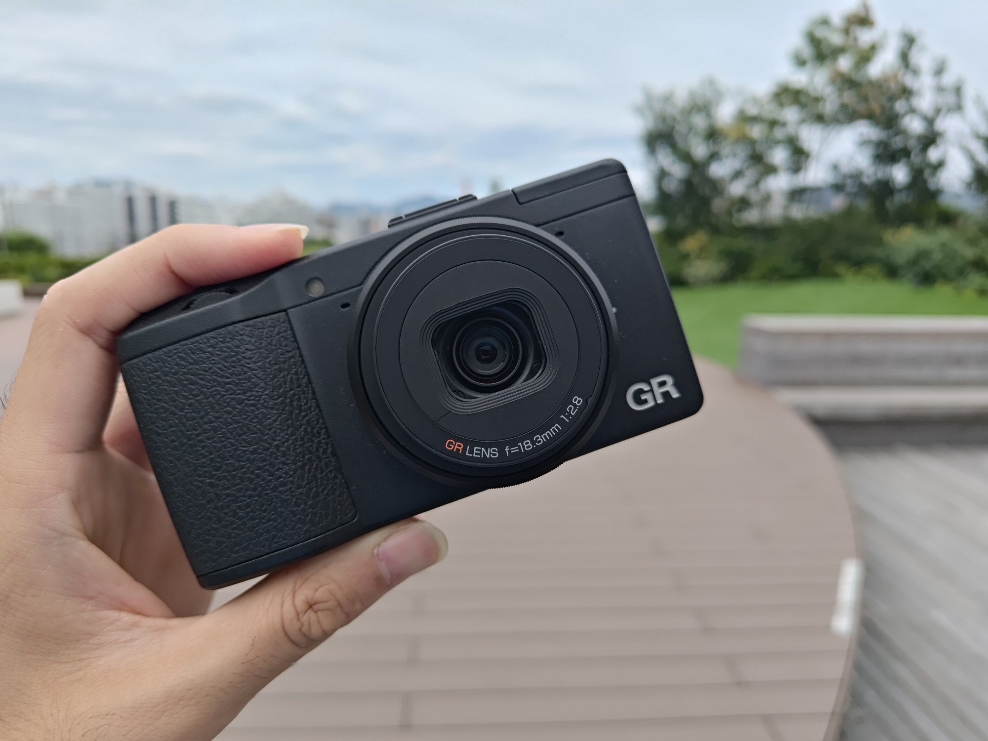 屋上デッキで、黒いRICOH GR（初代）を手に持った手元。レンズにGR LENS f=18.3mmの表記