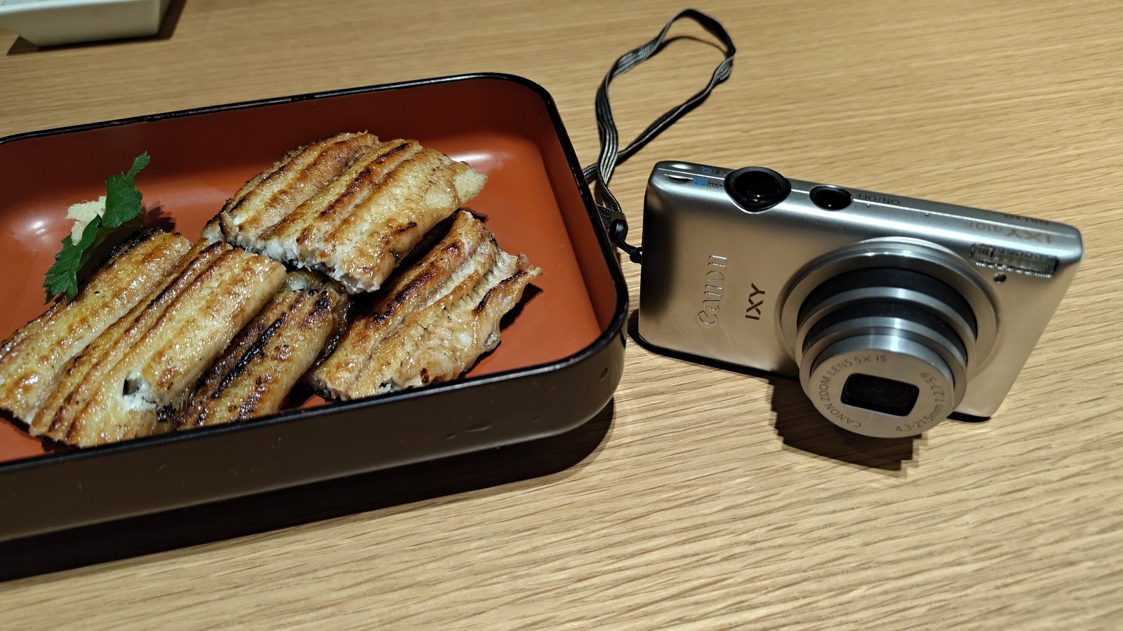 Canon IXY 410Fの本体と、赤い器に入った鰻の白焼きを並べて撮ったスマホ写真