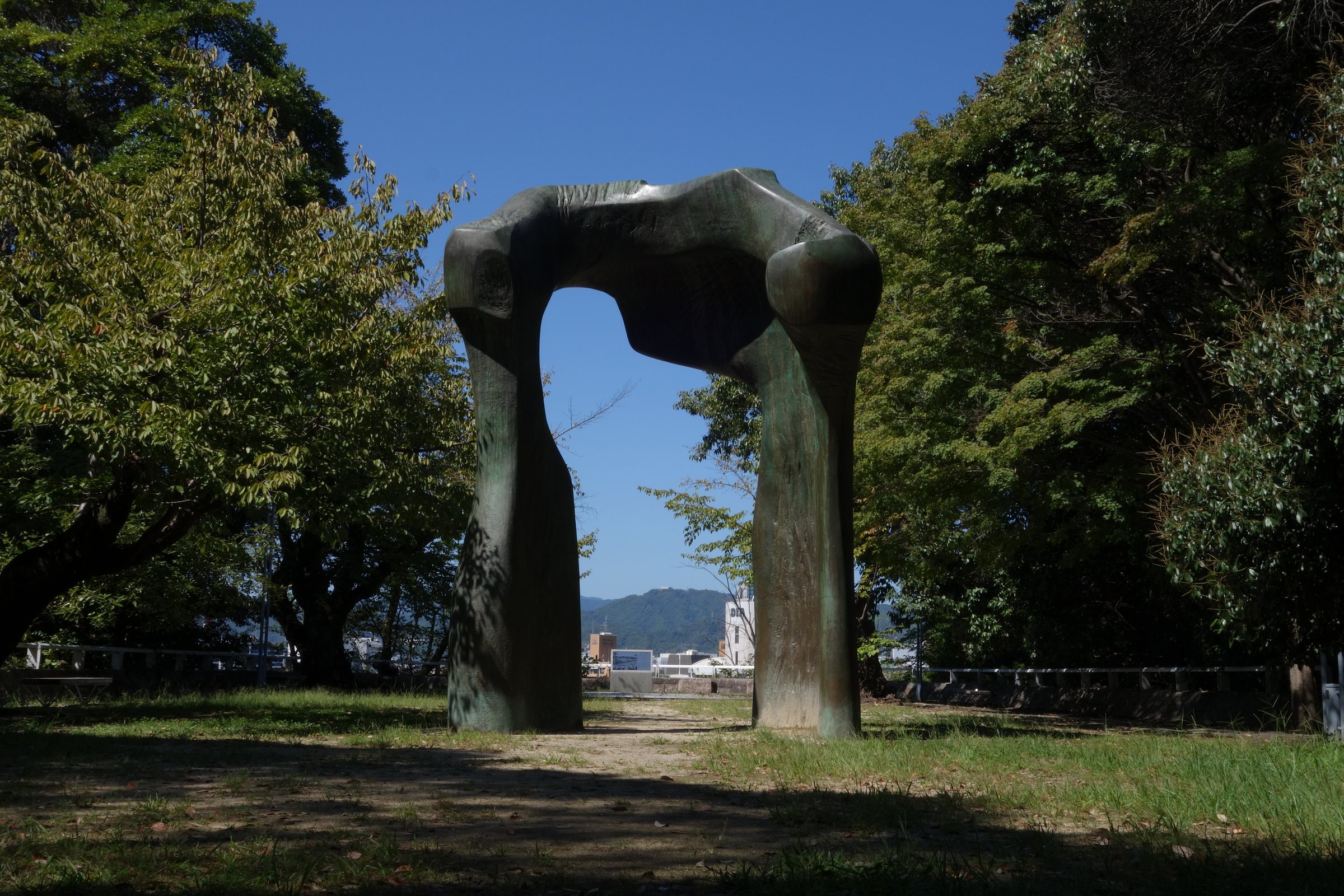青空の下、公園の広場に立つ、2本の脚の上を太い梁がつないだ門の形の大きな彫刻。彫刻の向こうに広島の街と山が見える