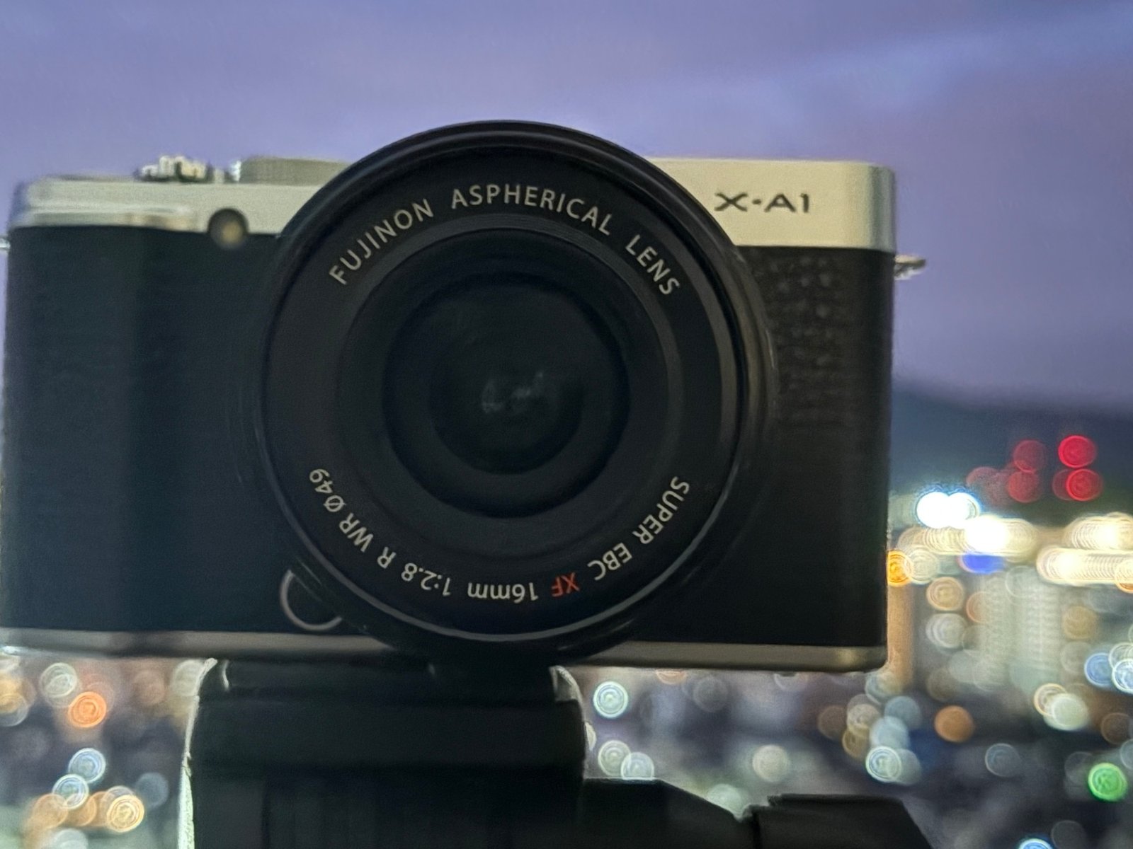 夕空とボケた街の明かりを背景に、三脚に載せたシルバーと黒のFUJIFILM X-A1と単焦点レンズの正面