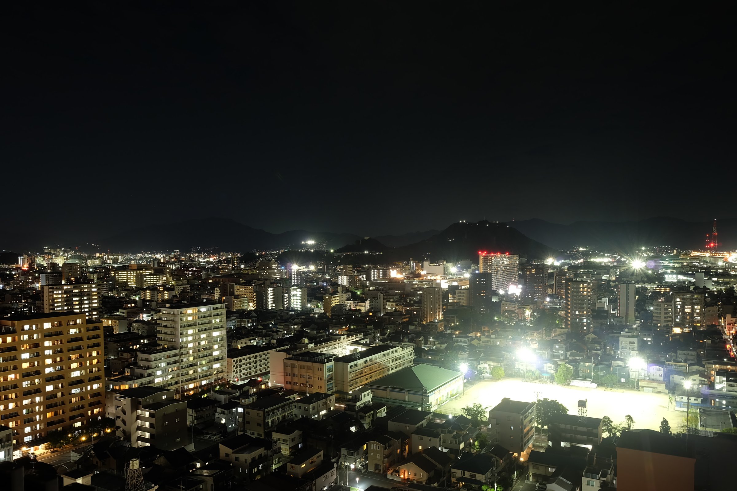 空が真っ黒で、マンションの窓の明かりがオレンジ色に光る夜景。右手前でグラウンドが白く光っている