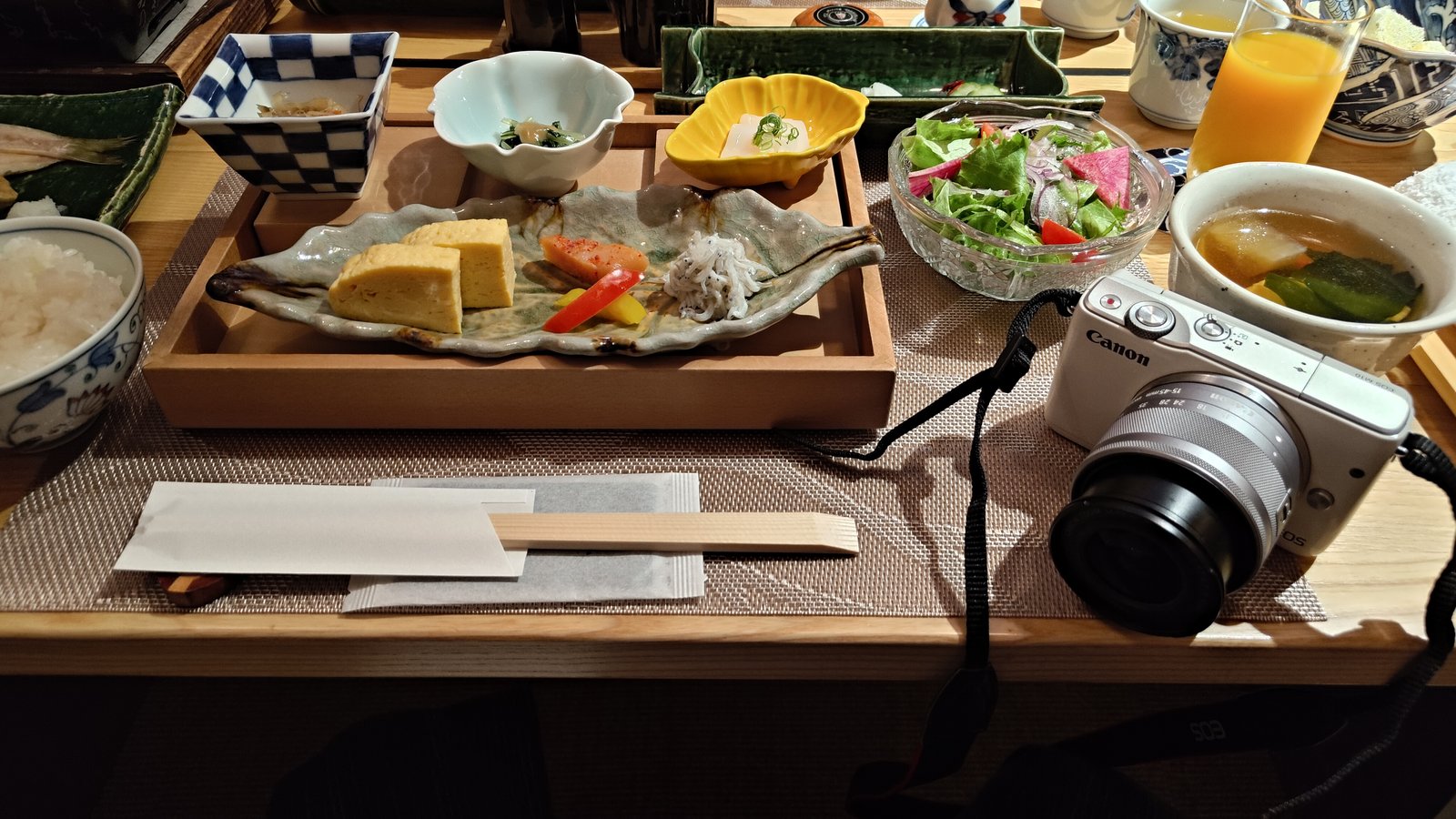 旅館の朝食の膳の右に置かれた、白いCanon EOS M10と標準ズームレンズ。左にだし巻き玉子の皿と汁椀が並ぶ