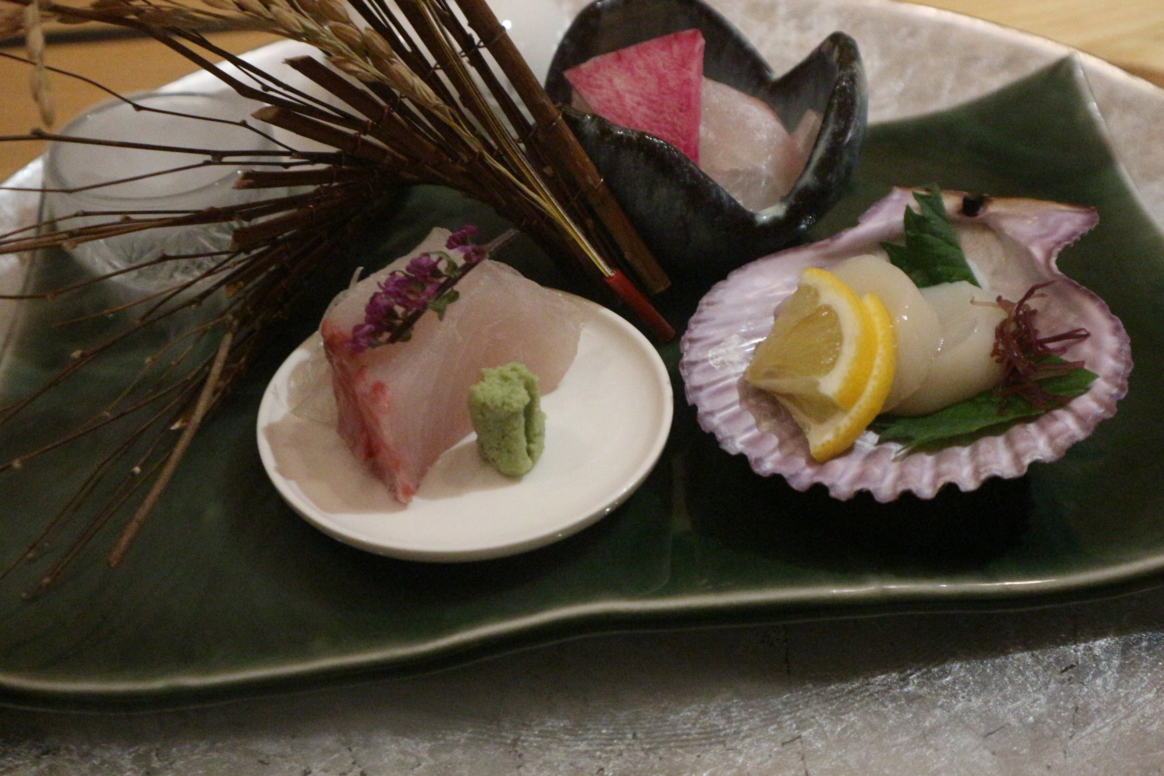 緑の皿にのったお造り。左の白い小皿に白身の刺身とわさび、右の貝殻にホタテとレモン。皿のまわりは暗い