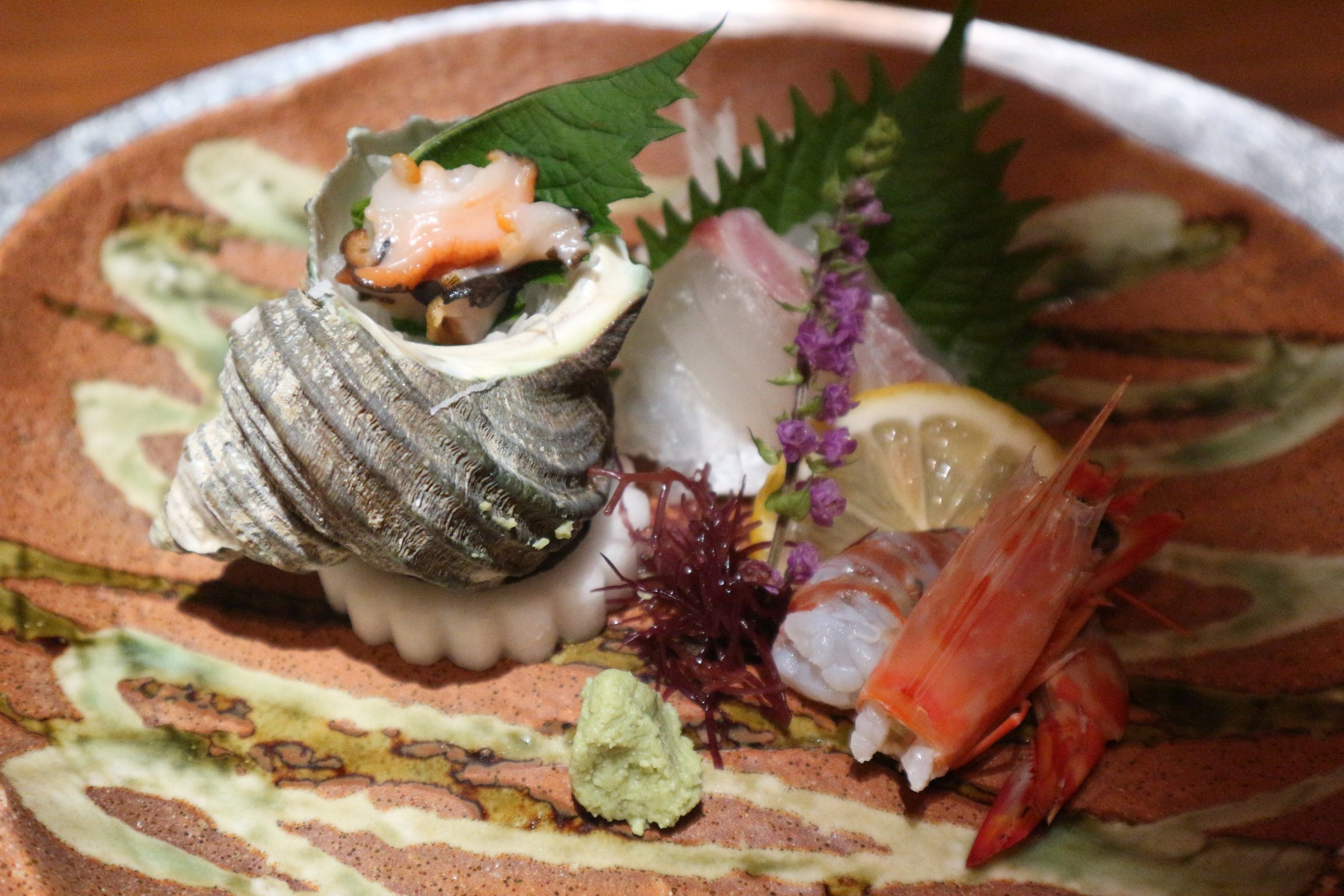 茶色の皿にのったお造り。左に巻貝、右に頭付きの海老とレモン、奥に白身の刺身と大葉が明るく写っている