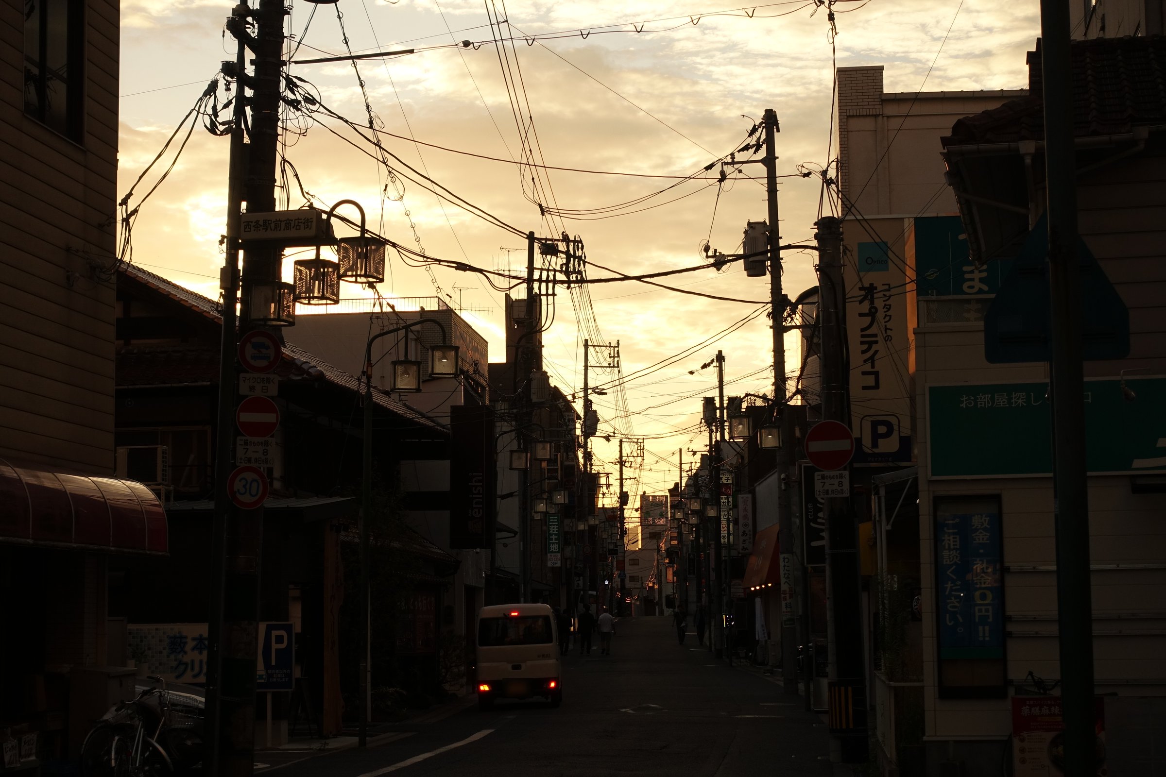 夕方の駅前商店街を、通りの奥へ向かって撮った夕景モードの写真。通りの奥の空が金色っぽく、電線と電柱は黒い影、左側の建物は暗い。通りの左寄りにクリーム色の軽バンが止まっている