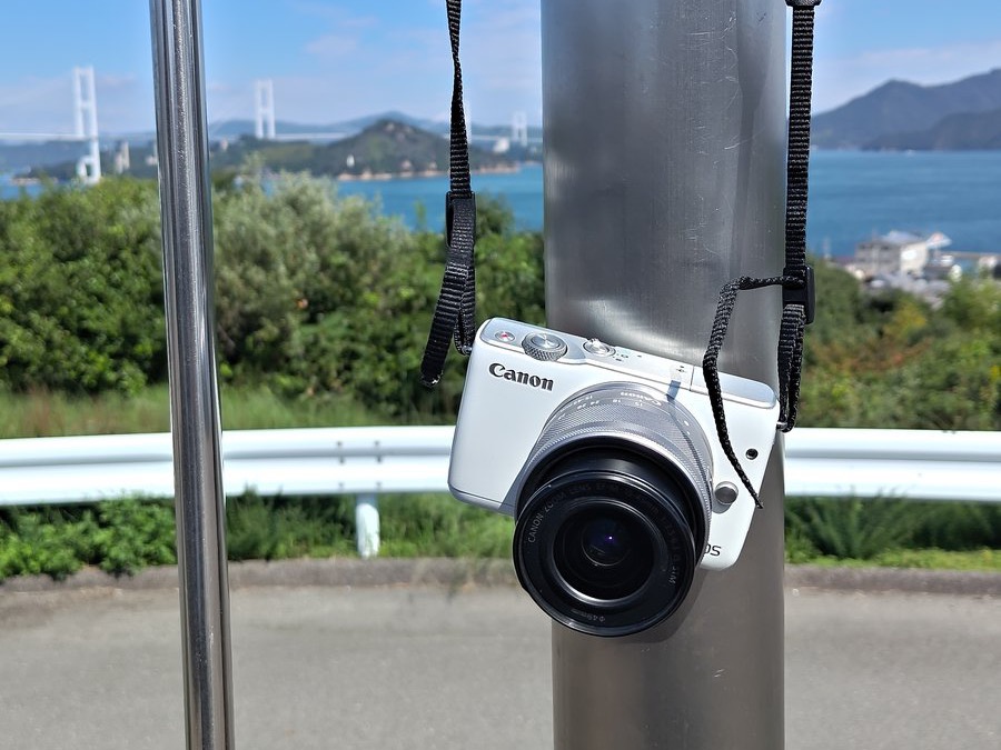 来島海峡大橋が見える場所で、金属のポールにストラップで掛けた白いCanon EOS M10